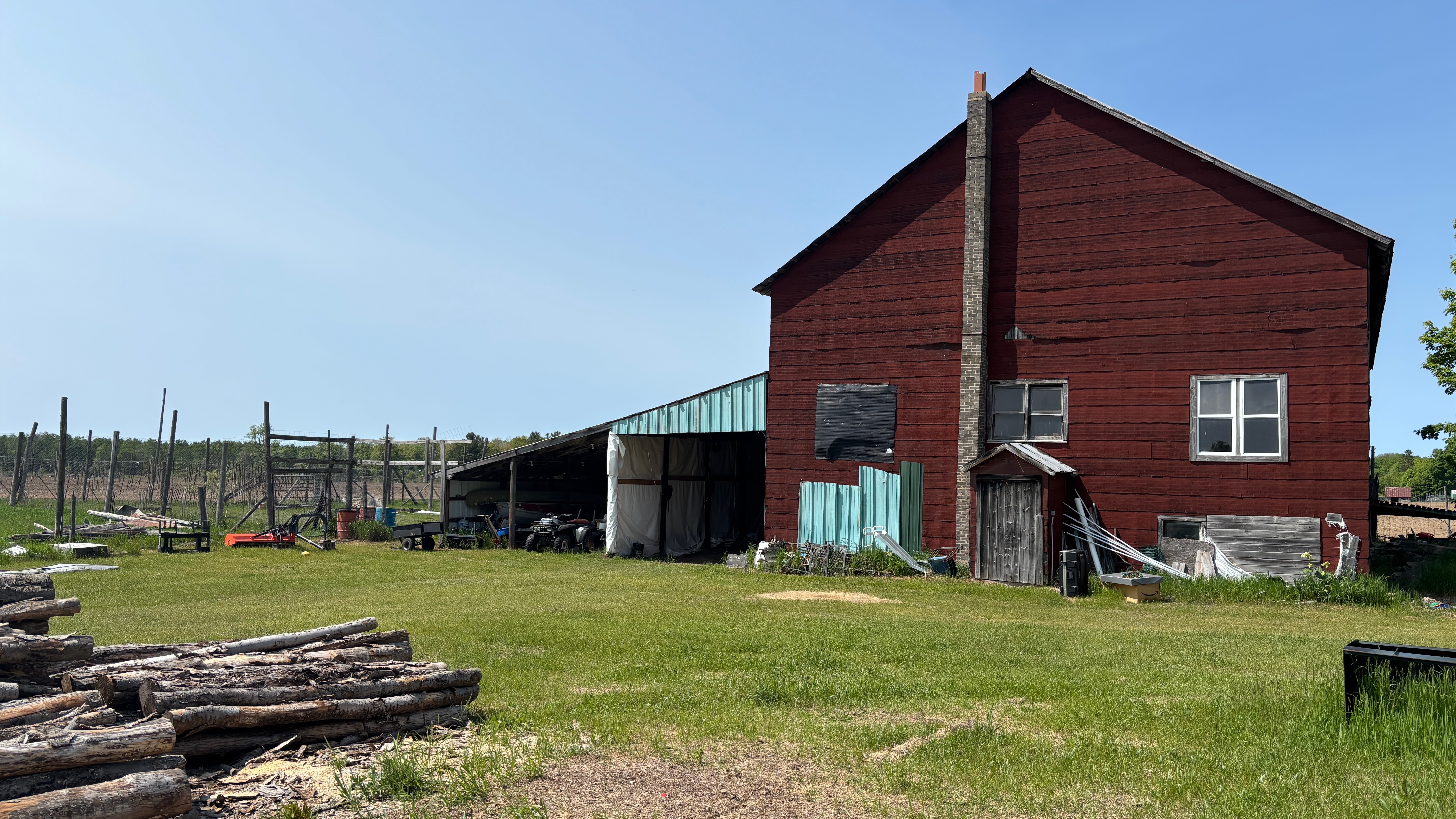 An old red barn on the right side of the photo. It has a very tall chimney on the outside of the barn. To the left some lean-tos built against the barn, and the left side of the photo is open clear sky. In the foreground some logs.