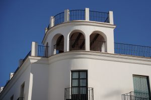 A close-up view of the upper section of a white building featuring a rounded architectural design with multiple archways and a balcony enclosed by black railings. The building displays a clean and modern aesthetic with minimal decorative features.
