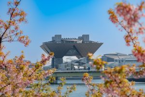 Tokyo Big Sight stands tall under a blue sky, framed by cherry blossoms in pink and green, with Ariake Pier Bridge and Tokyo Bay in the foreground.