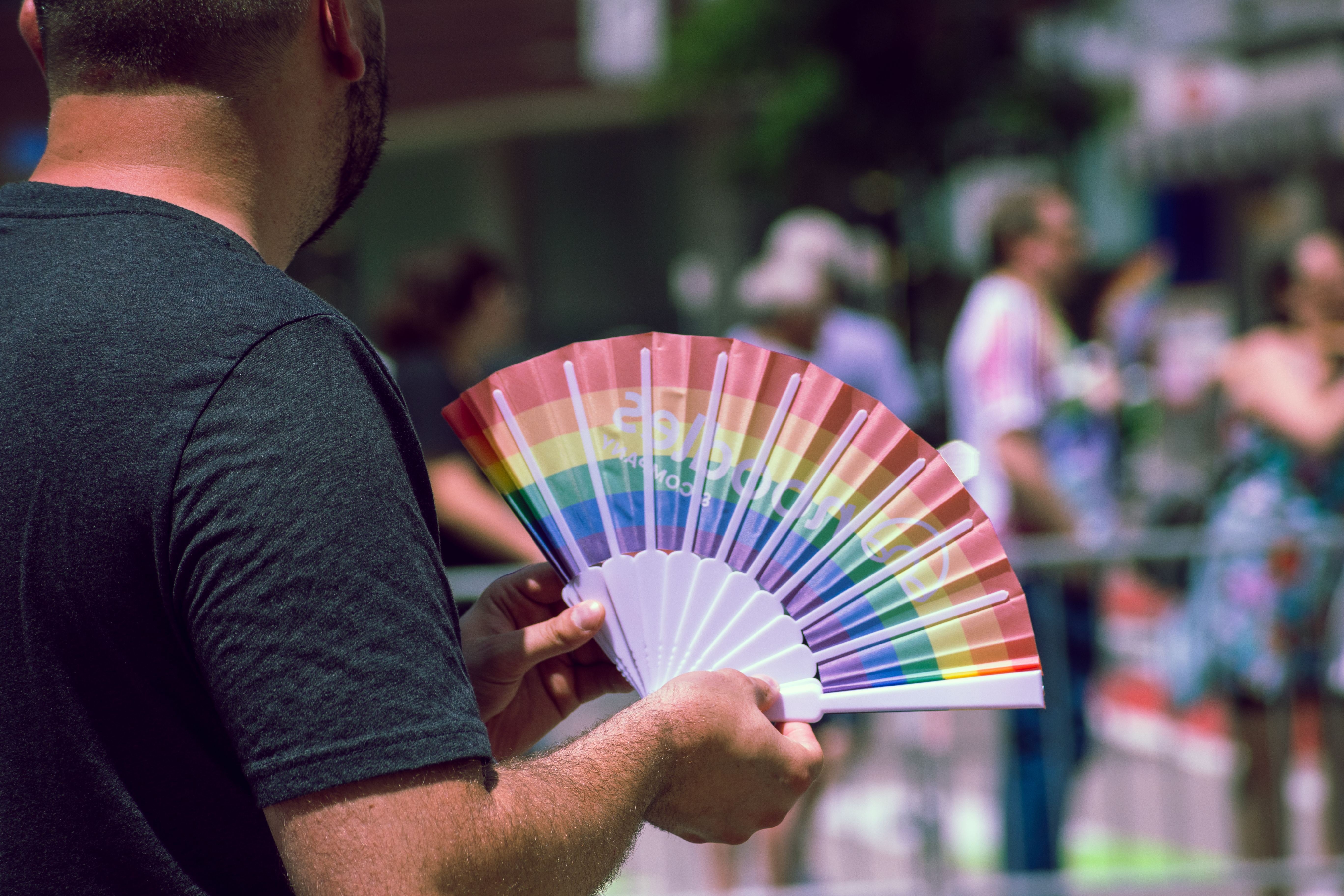 A person in a dark t-shirt holds an open rainbow fan at a Pride event. Blurred figures in colorful clothing and barricades are seen in the background on a sunny city street.