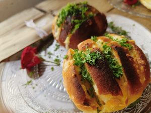 Two golden-brown bread rolls, scored and filled with melted cheese, rest on a clear glass plate. They’re topped with fresh parsley. A red rose lies beside them, adding a romantic touch. The background shows a wooden table and a softly blurred parcel tied with string.