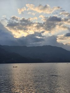 A serene lake scene during sunset, with soft waves reflecting the colorful sky filled with clouds. The silhouette of distant mountains lines the horizon, while several small boats can be seen gently floating on the water.
