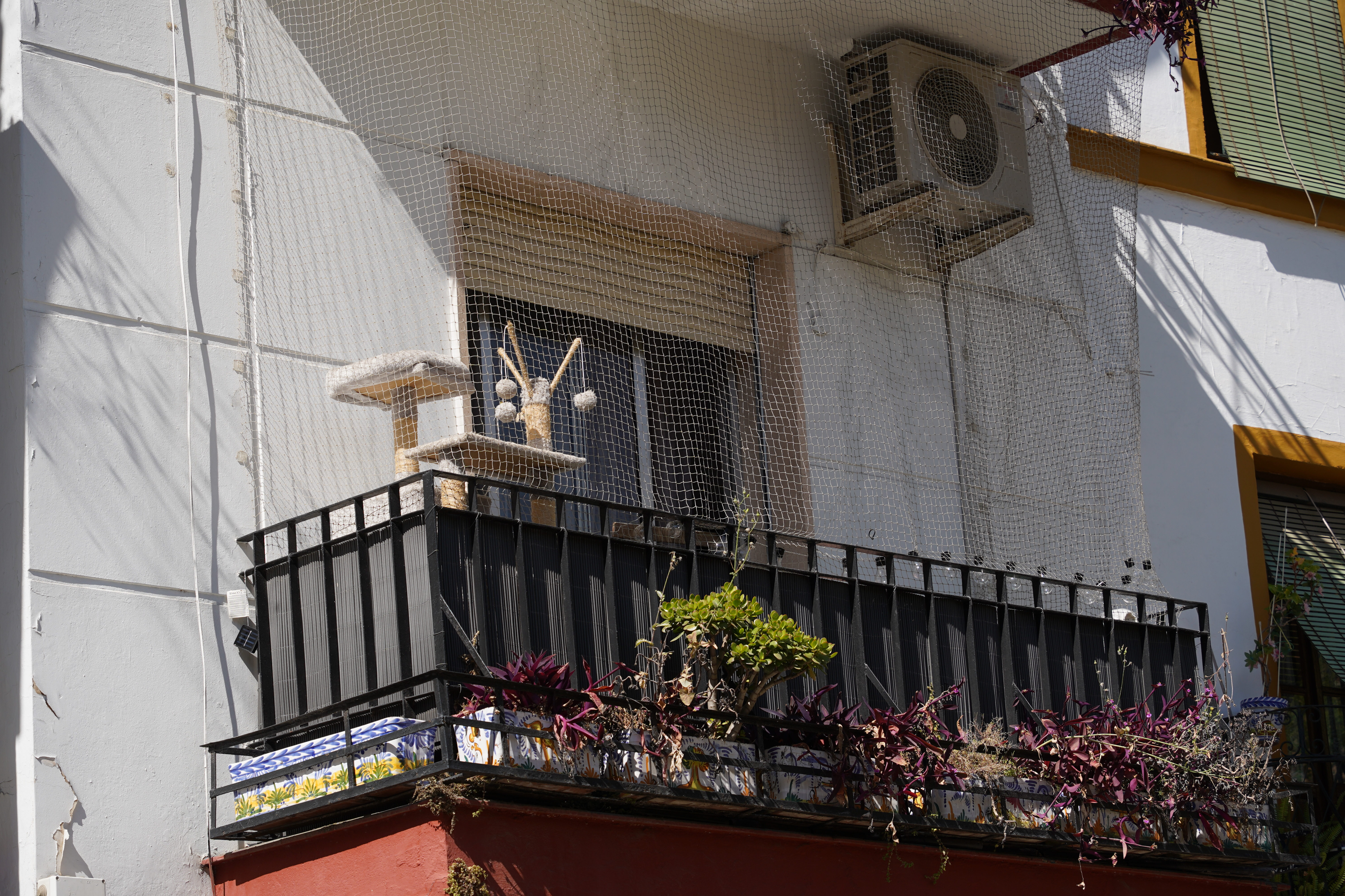 A balcony with a black railing and decorative potted plants. A cat tree with a small platform and dangling toys is positioned on the balcony. There is a fine netting is stretched across the area, to prevent pets from escaping.
