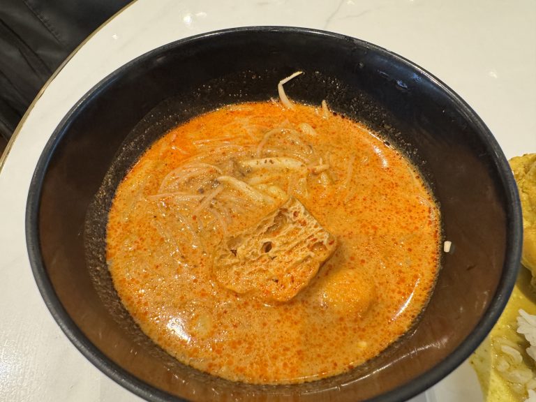 A bowl of spicy noodle soup with tofu and bean sprouts in orange broth.