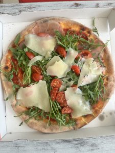 A freshly baked Italian pizza topped with arugula, cherry tomatoes, thin slices of prosciutto, and generous shavings of parmesan, served in an open takeaway box.