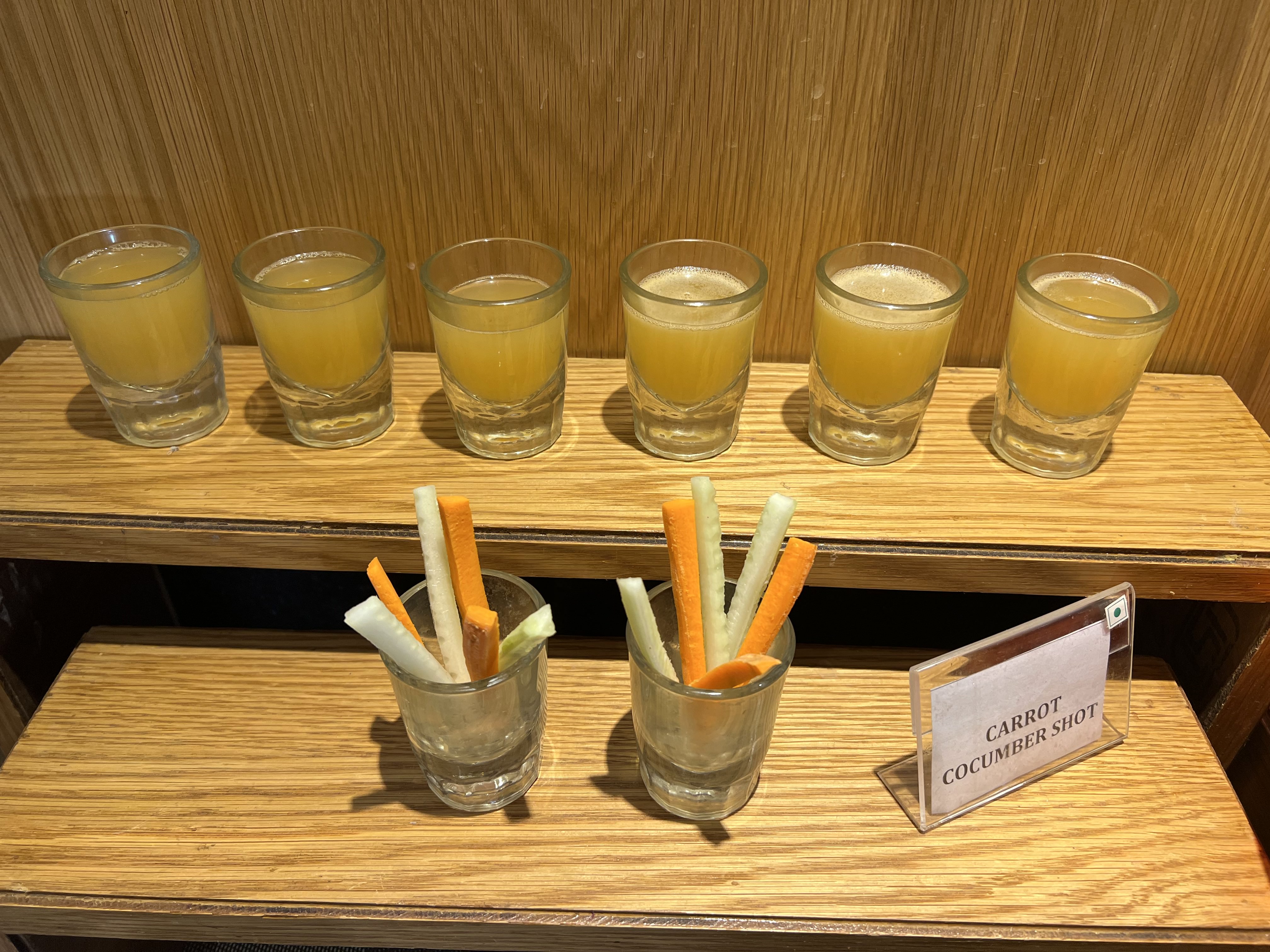 A wooden shelf displays eight small glasses filled with a yellow drink, arranged in two rows. Below the glasses, a small container holds sticks of carrot and cucumber, with a label reading "CARROT CUCUMBER SHOT" beside it.