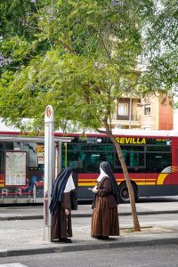 Two nuns dressed in traditional dark brown clothing and white head coverings stand near a bus stop. One woman is holding a piece of paper and appears to be talking to the other.
