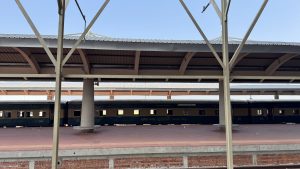 A train is seen at a railway platform, partially obscured by structural supports.