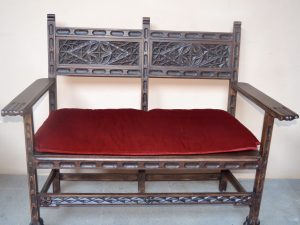 A vintage or antique wooden settee featuring intricate carved details on its backrest and claw-footed legs. A vibrant red velvet cushion provides seating on the dark-stained wooden frame. Captured from Tenerife, Spain. 