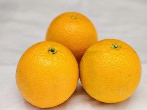 Three whole oranges are arranged on a light-colored surface. Two oranges are positioned in the foreground, while one is slightly behind and centered. It was captured in Kozhikode, Kerala.