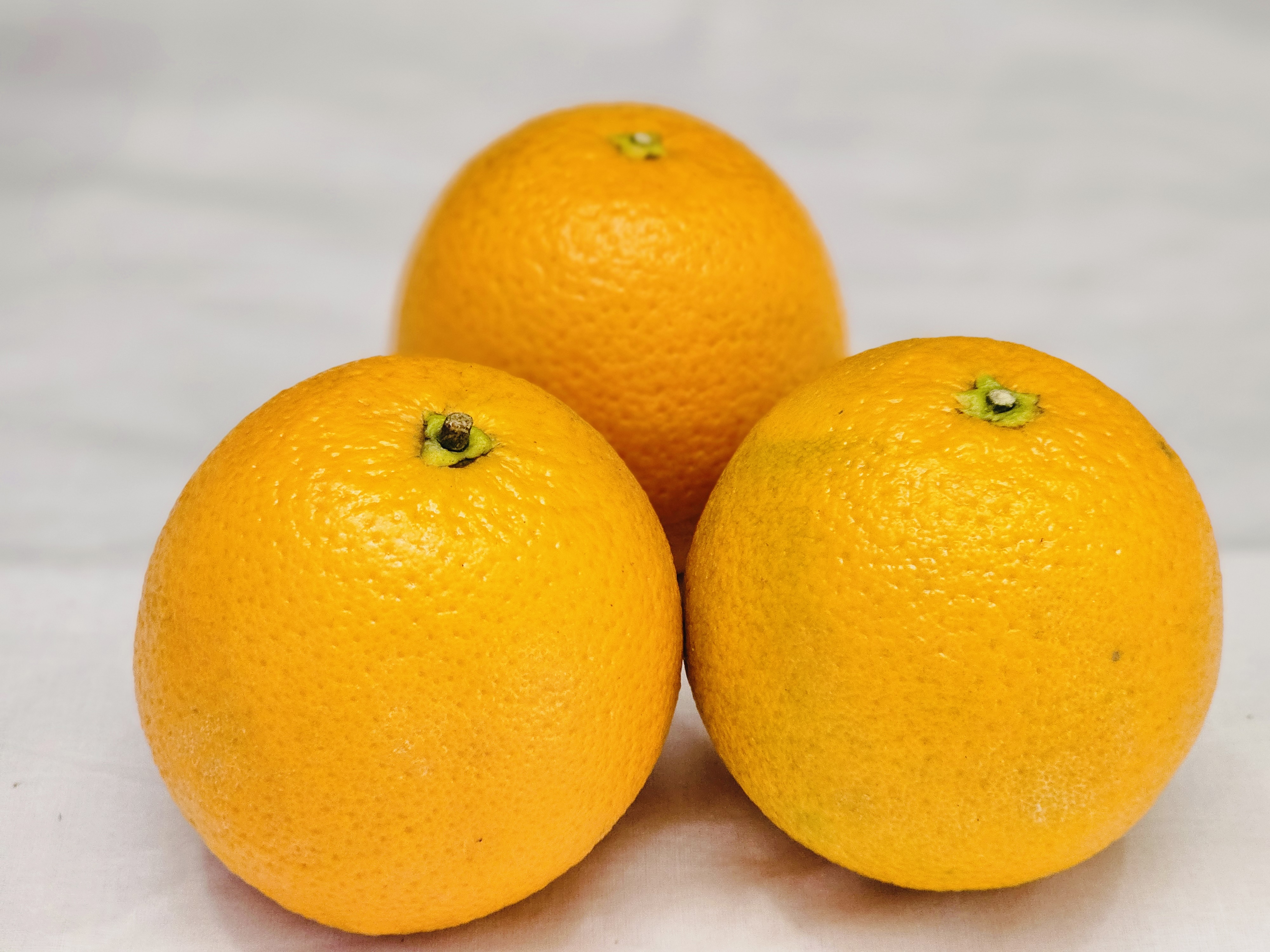 Three whole oranges are arranged on a light-colored surface. Two oranges are positioned in the foreground, while one is slightly behind and centered. It was captured in Kozhikode, Kerala.