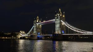 A night view of Tower Bridge in London, illuminated with vibrant lights against a dark sky. The bridge spans the River Thames, reflecting light on the water's surface. Nearby buildings and a distant skyline are visible, adding to the urban ambiance.
