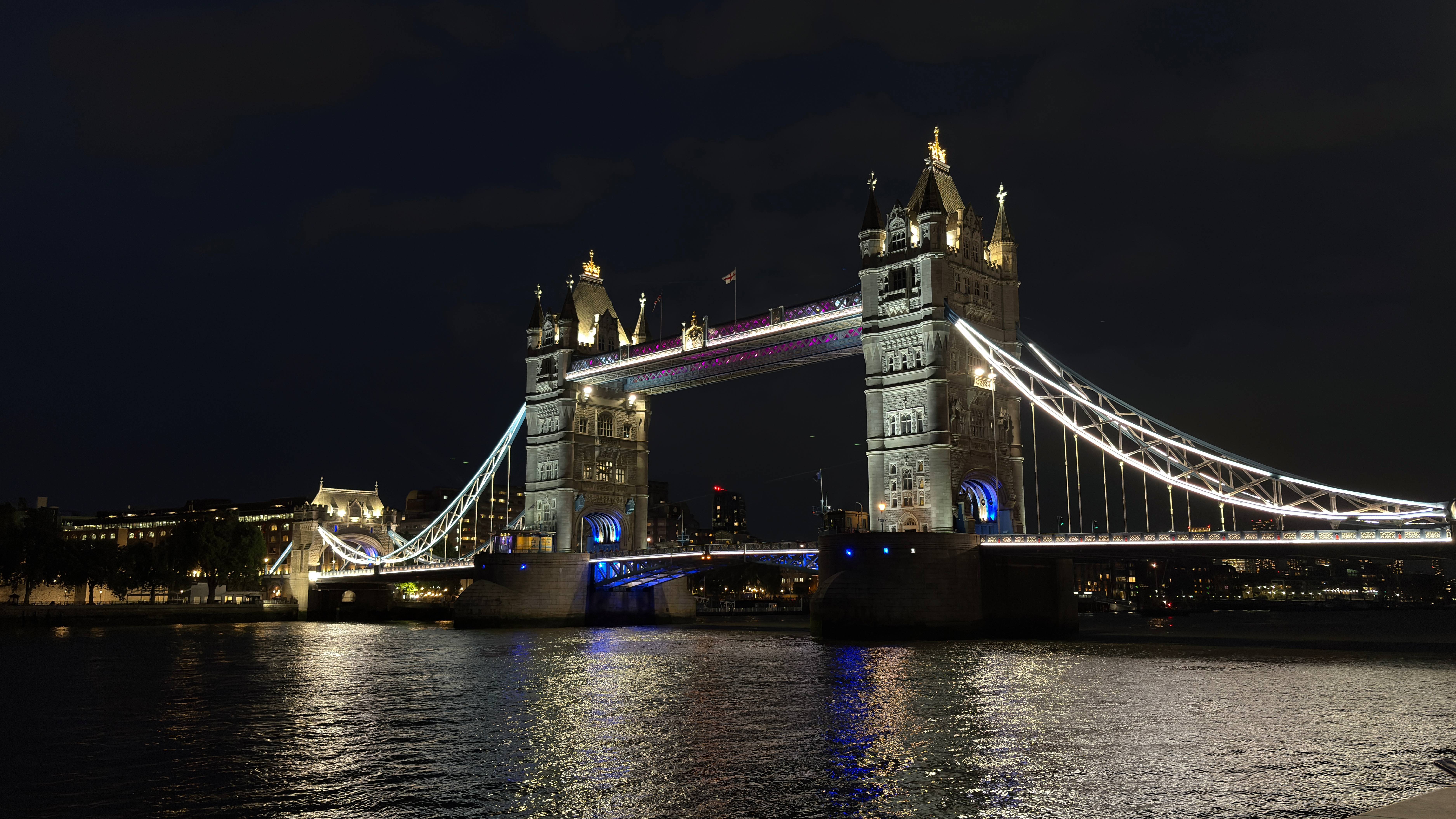 A night view of Tower Bridge in London, illuminated with vibrant lights against a dark sky. The bridge spans the River Thames, reflecting light on the water's surface. Nearby buildings and a distant skyline are visible, adding to the urban ambiance.
