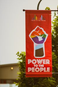 A red banner reads "Twin Cities Pride" at the top with a rainbow bridge logo and a rainbow fist in the center and the text "Power to the People" at the bottom. In the background is a part of a tree and a low building.