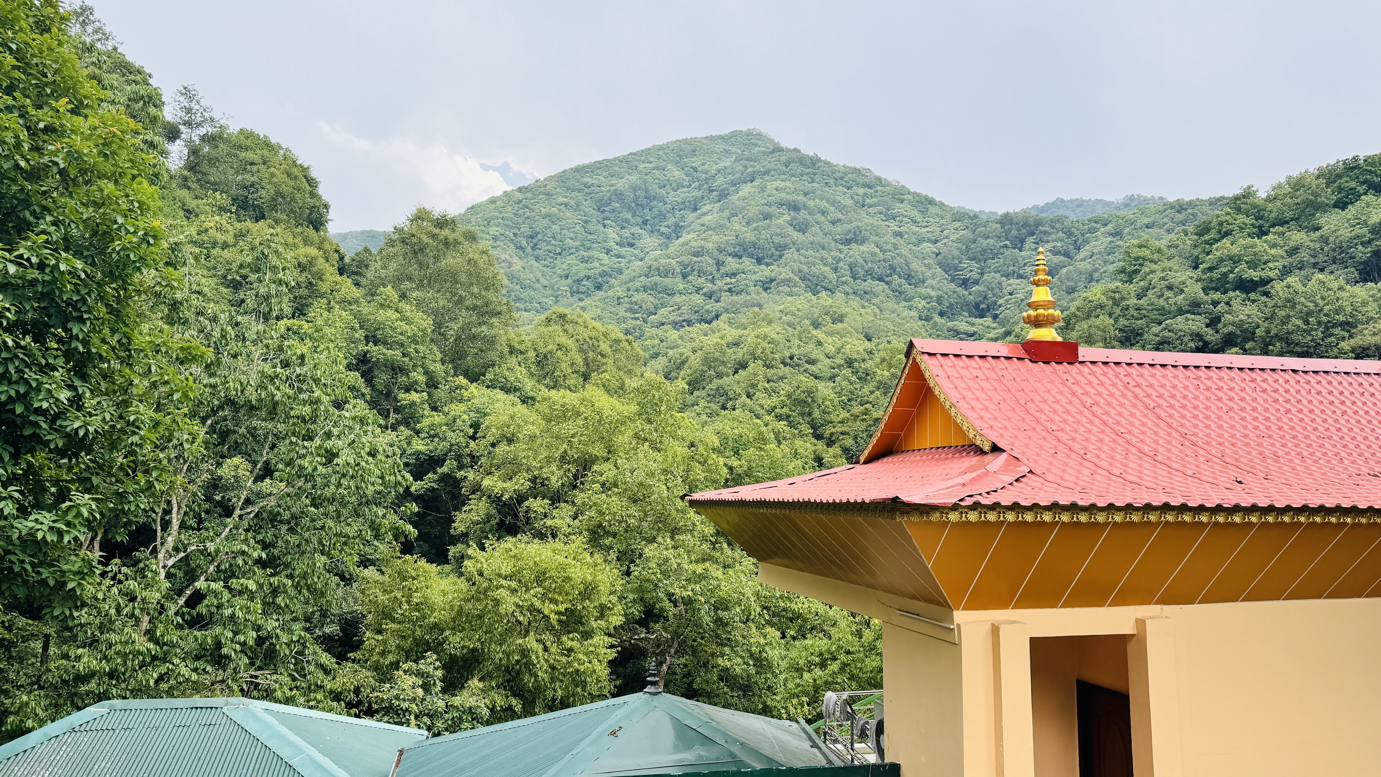 A view of lush green mountains in the background, with a temple featuring a vibrant red and gold roof in the foreground.