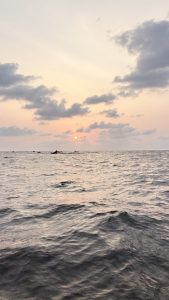 A scenic view of the ocean at sunset, with the sun partially visible on the horizon surrounded by soft, fluffy clouds. 