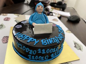 A chocolate birthday cake with a glossy black frosting features a whimsical figurine of a person in a blue hoodie sitting in front of a laptop.
