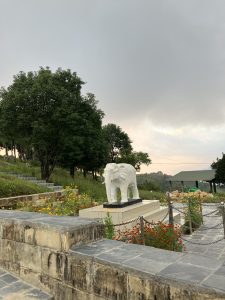 A white stone elephant statue stands on a pedestal surrounded by vibrant flowers in a landscaped garden.