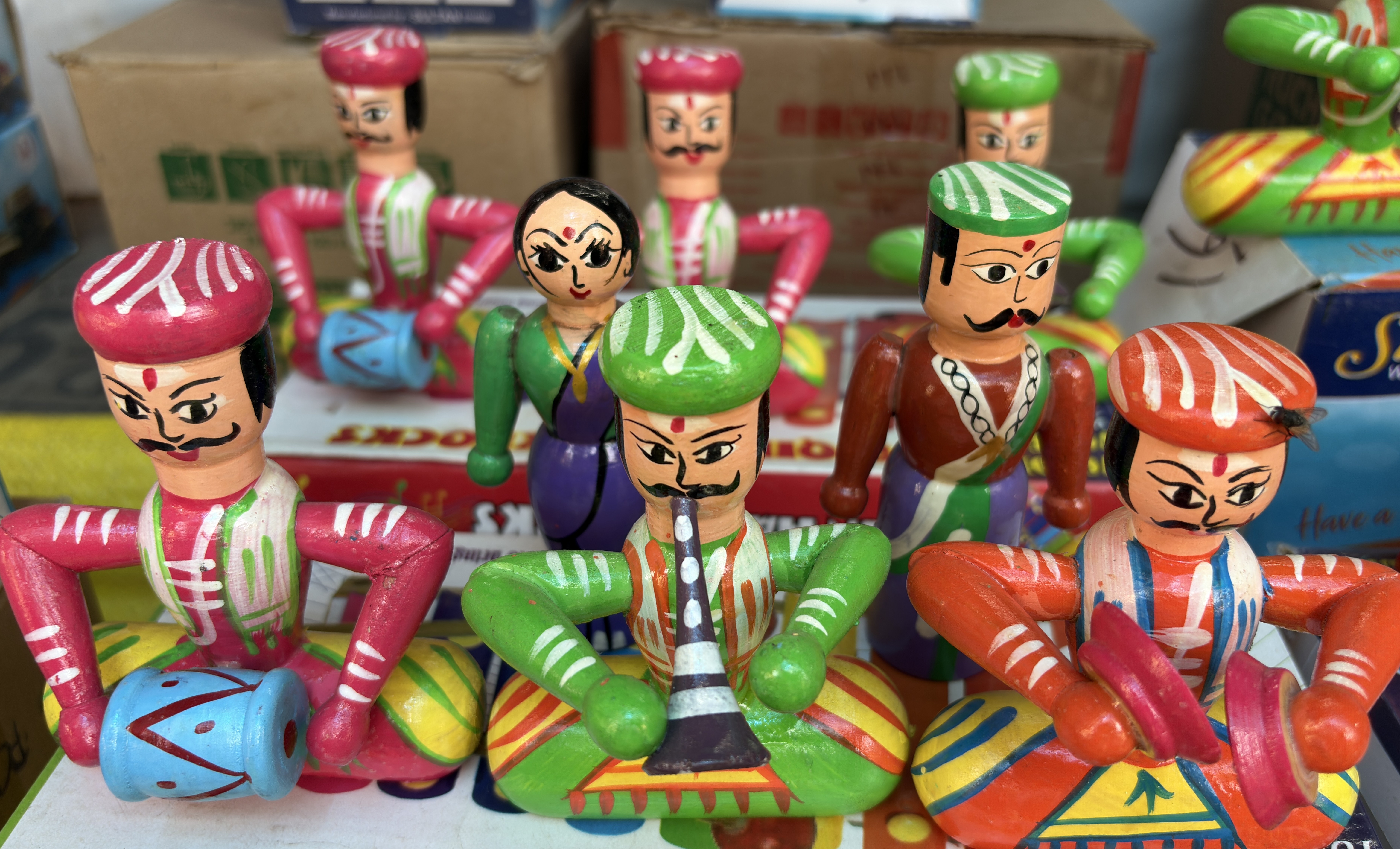 A collection of colorful wooden dolls depicting traditional Indian musicians and dancers. Each figure is hand-painted with vibrant clothing and expressive facial features, holding musical instruments like drums, cymbals, and a shehnai.