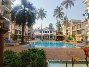 A clear turquoise pool sits in a peaceful courtyard, surrounded by tall white residential buildings with balconies. Palm trees and lush greenery add tropical charm. Lounge chairs line the deck, and a lifebuoy hangs nearby. A small white gazebo and blue accents complete the serene setting.
