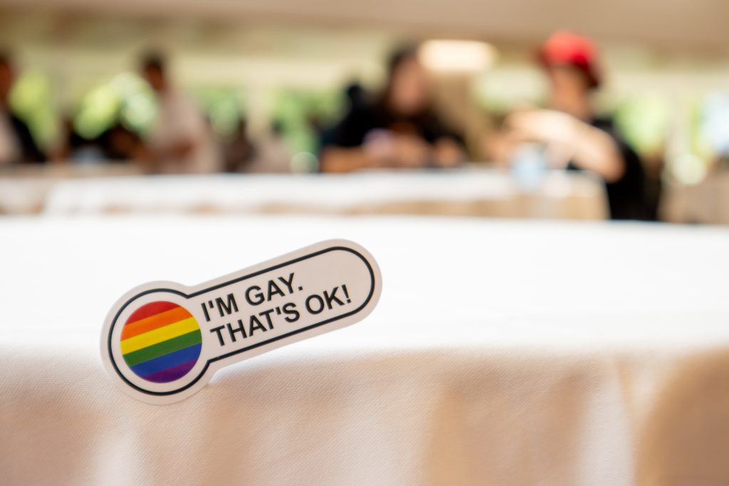 A sticker on a white tablecloth reads ‘I’M GAY. THAT’S OK!’ with a rainbow pride circle. The background is softly blurred, showing people gathered in conversation.