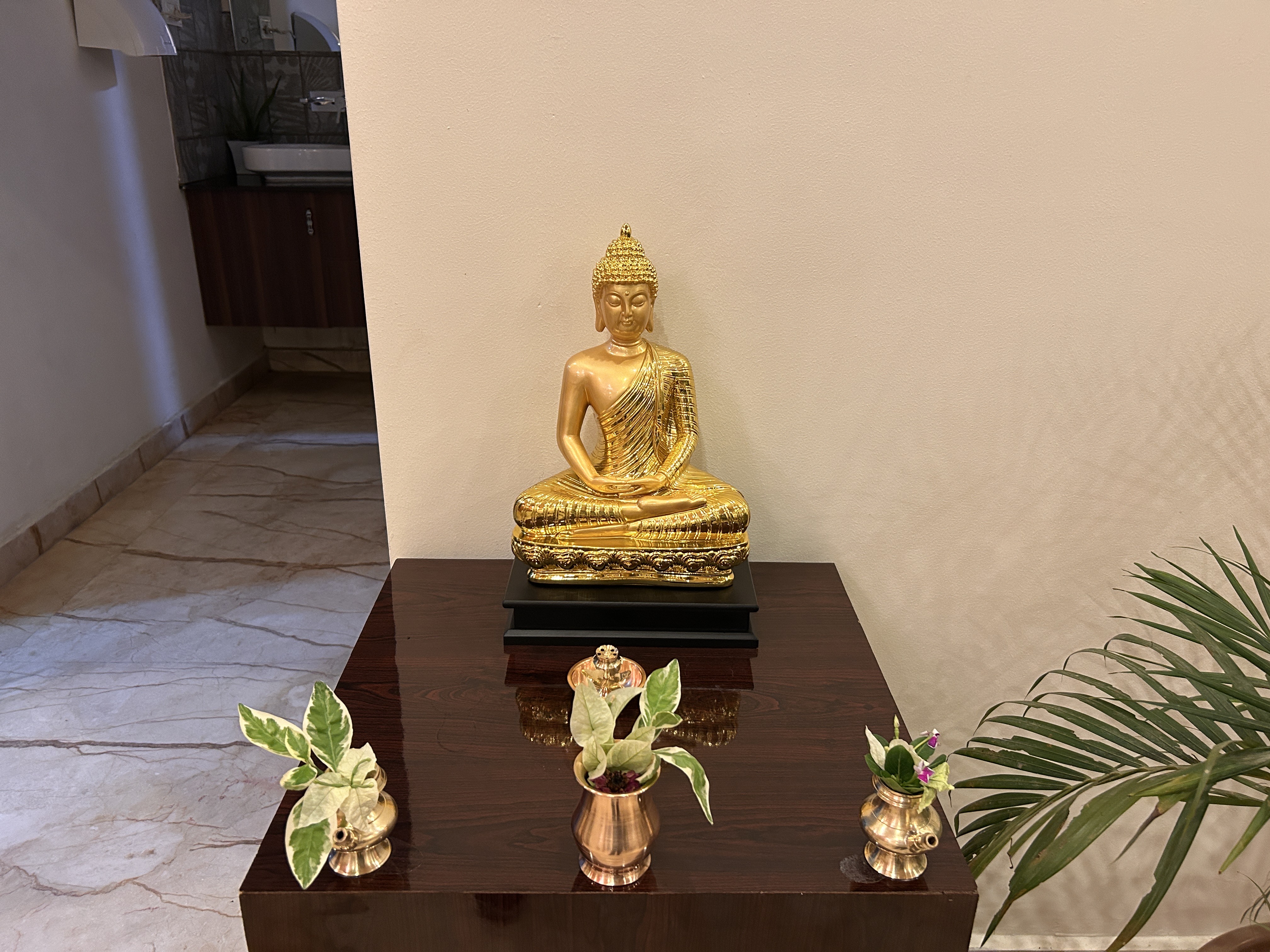 A golden statue of Buddha is placed on a dark wooden table. In front of the statue, there are three small decorative pots with green plants, and a brass pot is positioned in the center.