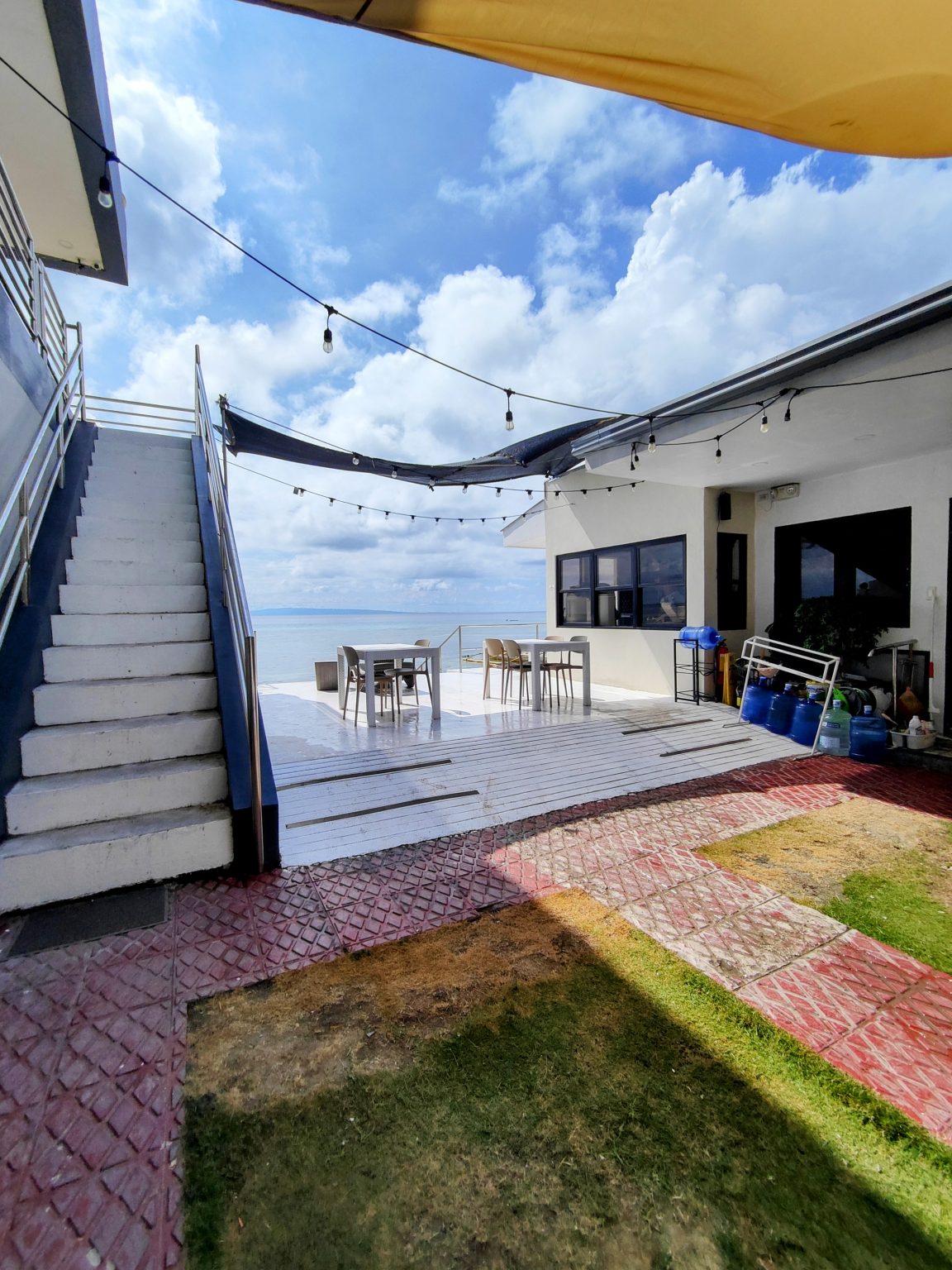 A bright and breezy coastal view from a beachside property in Oslob, Philippines. Featuring a clean outdoor seating area, string lights, and ocean views under a blue sky with fluffy clouds.
