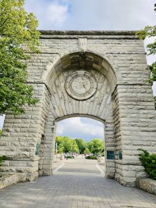 Adams Arch, a grand stone structure with a circular emblem, frames a view of parked cars and lush trees near Niagara Falls.