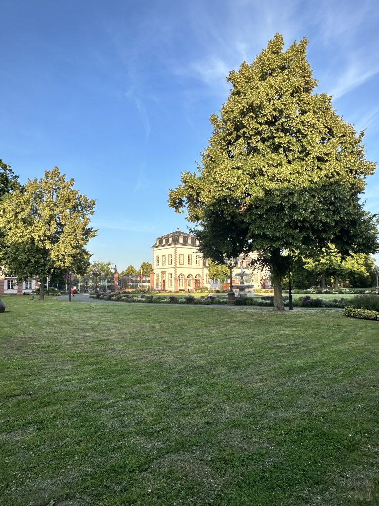A beautifully manicured park with lush green grass and tall trees surrounds a grand historic building bathed in golden sunlight, under a vibrant blue sky.