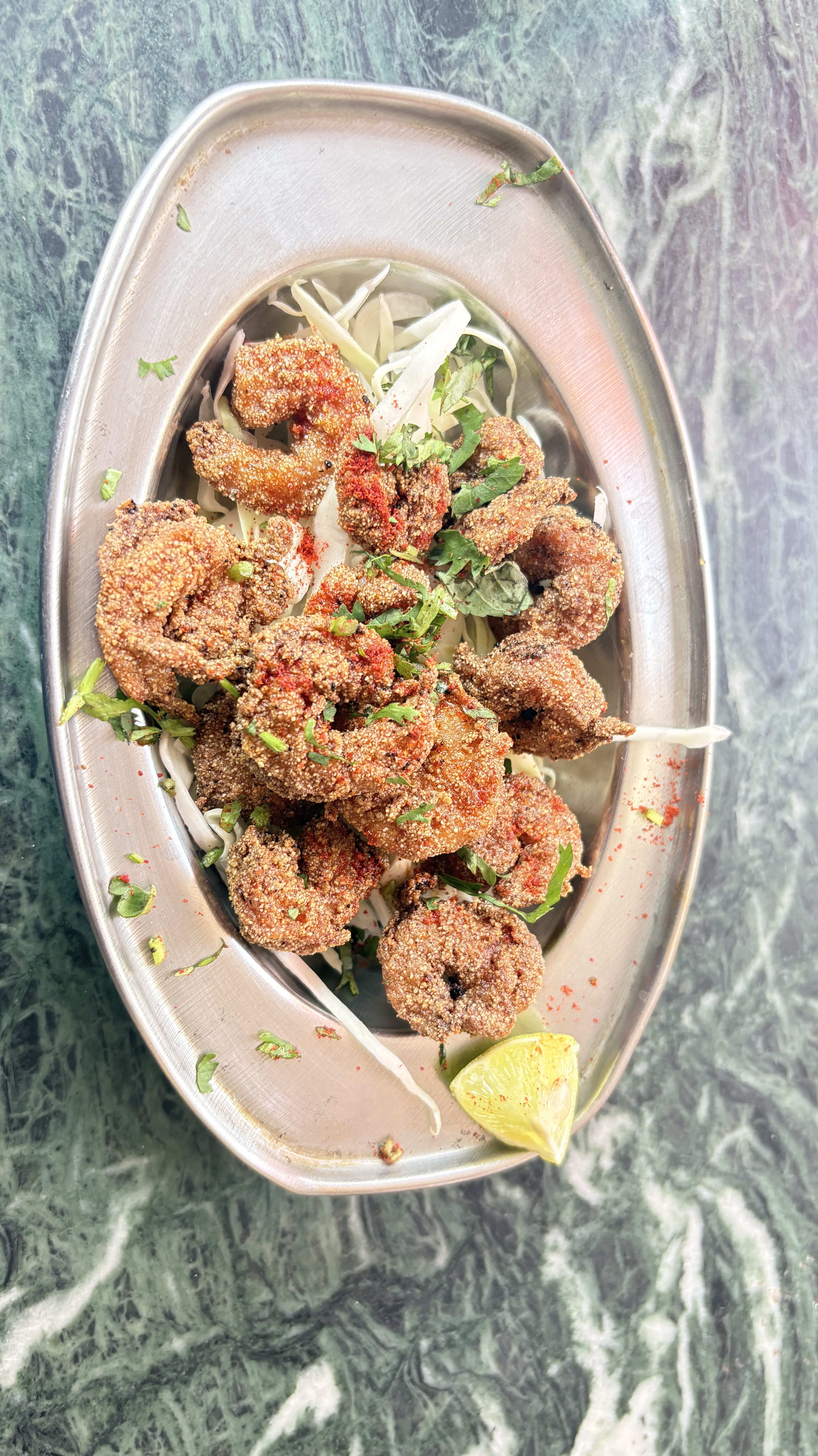A silver platter holds a serving of fried shrimp, garnished with chopped cilantro and a sprinkle of red spice.