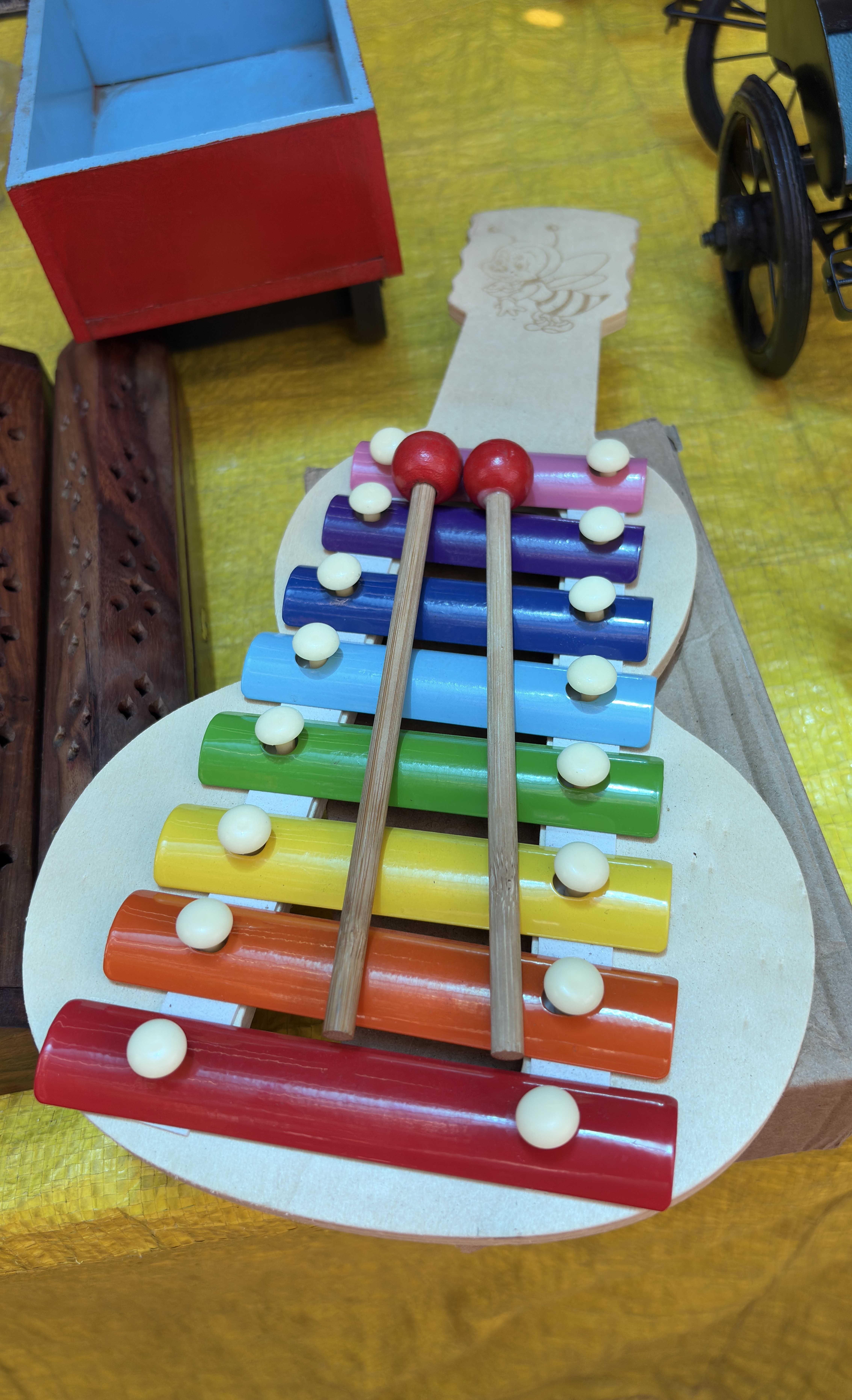 A colorful xylophone toy shaped like a guitar, with eight rainbow-colored metal bars and two wooden mallets placed on top. The toy rests on a yellow surface, surrounded by other wooden toys including a red and blue cart and a miniature tricycle.