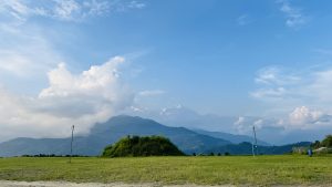 A panoramic view of a serene landscape featuring lush green fields in the foreground, with rolling hills and mountains in the background.