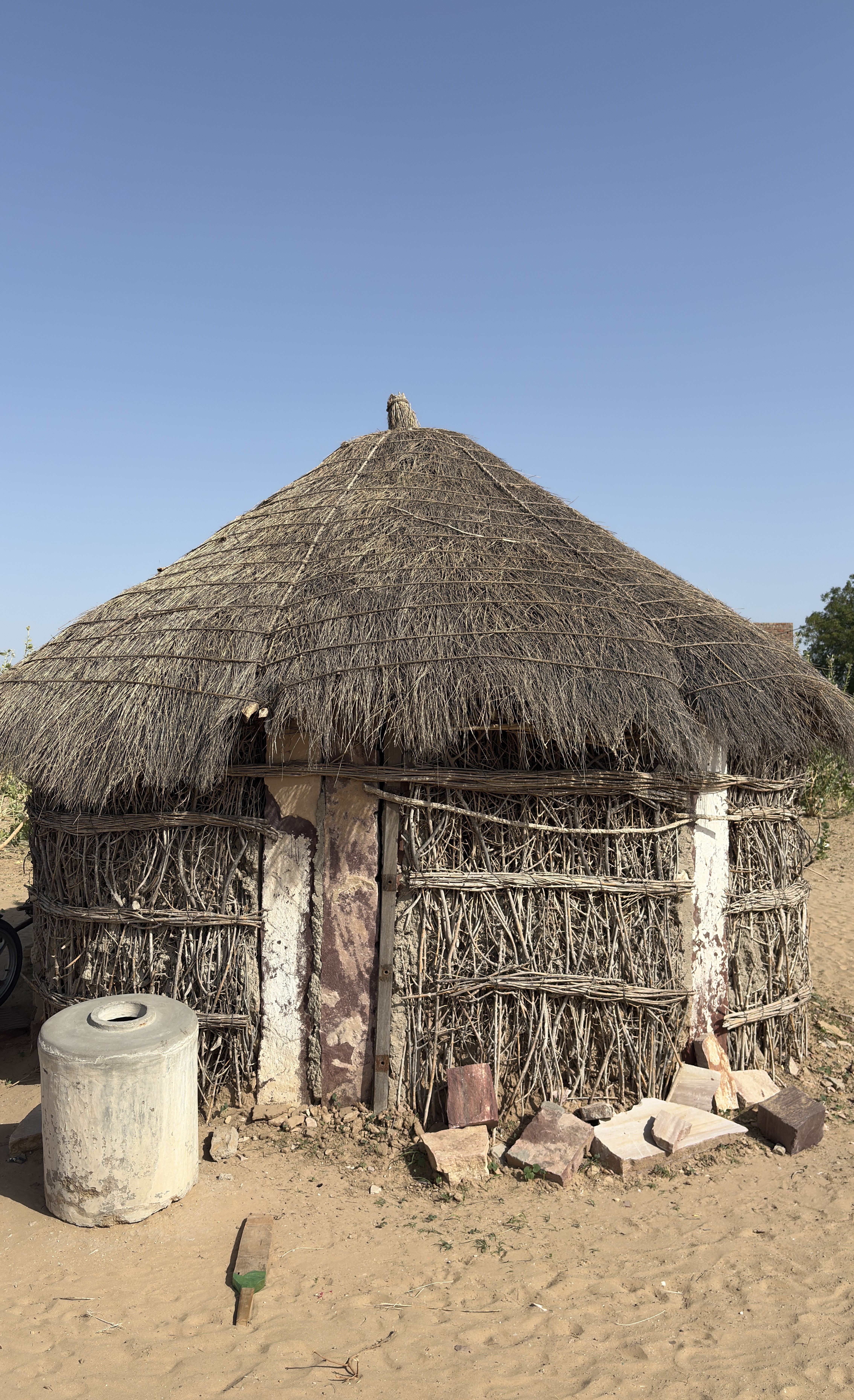 A traditional round hut constructed with woven branches and thatched roofing, set against a clear blue sky.