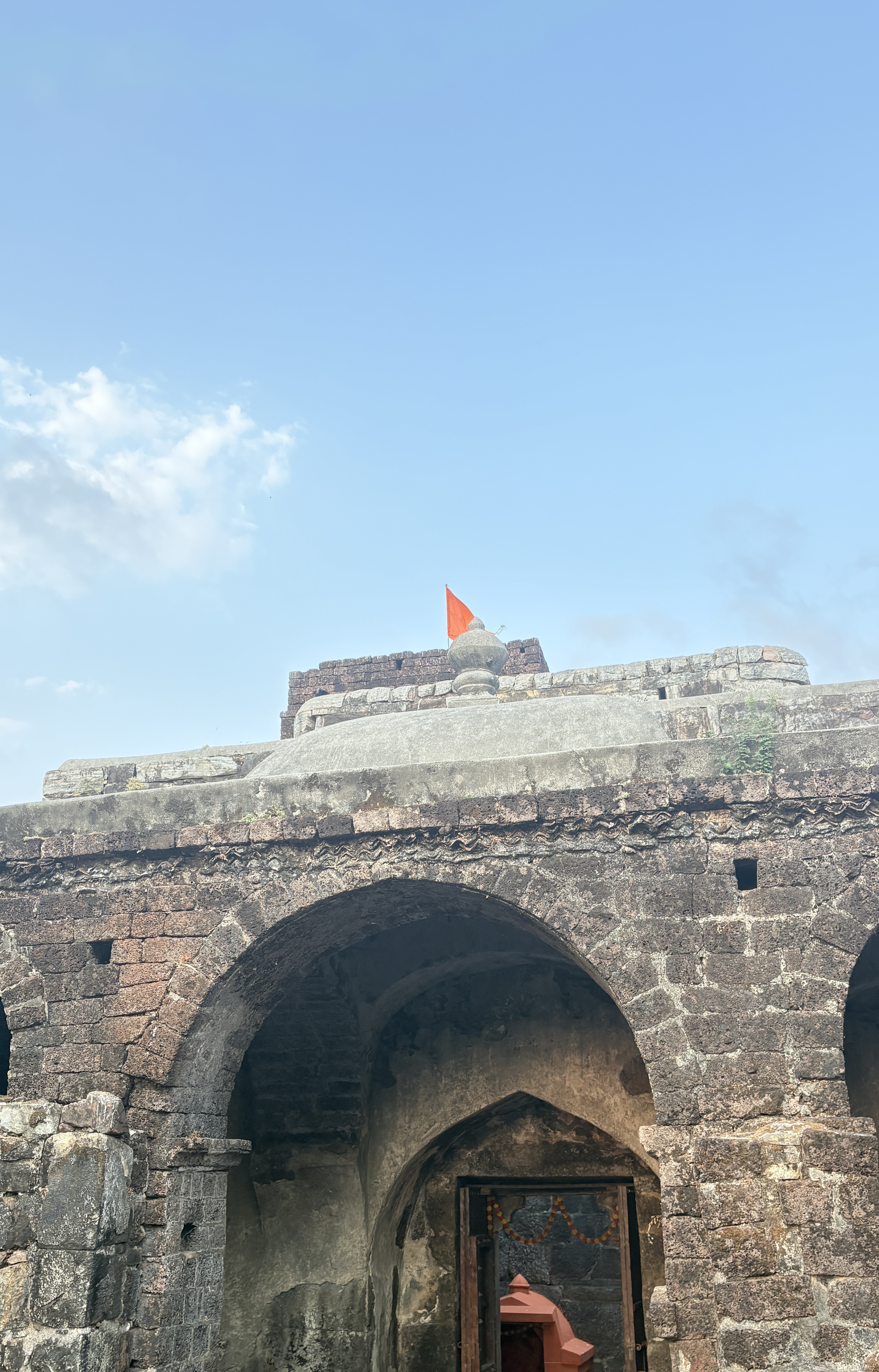An ancient stone structure featuring arches and a flat rooftop, atop which there is an orange flag fluttering against a blue sky with some clouds.