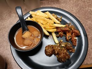 A black plate features a variety of foods: a bowl of brown sauce with small fried dumplings, crispy golden French fries sprinkled with seasoning, and two types of fried items.