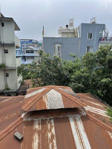 A view from above showing a rusty, corrugated metal roof in the foreground with a distinctive pointed structure. In the background, several buildings are visible, including a bright blue structure and a gray building with windows and a rooftop terrace.