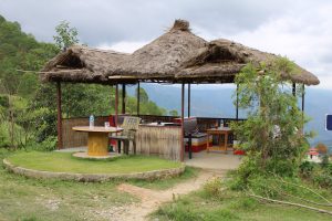 A thatched-roof gazebo in a scenic outdoor setting, featuring a round table with a yellow pedestal and a plastic chair. Nearby, there are two water bottles and a glass on the table. The gazebo has bamboo fencing, and there are comfortable seating arrangements inside