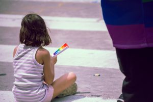 A young child in a striped tank top sits on a crosswalk holding a small rainbow flag during a Pride parade. Their gaze is directed toward the street as a person draped in a rainbow-colored garment stands nearby. 
