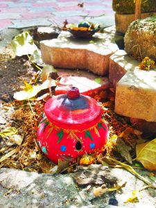 A hand-painted red clay Garbo pot used in Navratri rituals, placed among dry leaves with a lit oil lamp on stone bricks behind it.