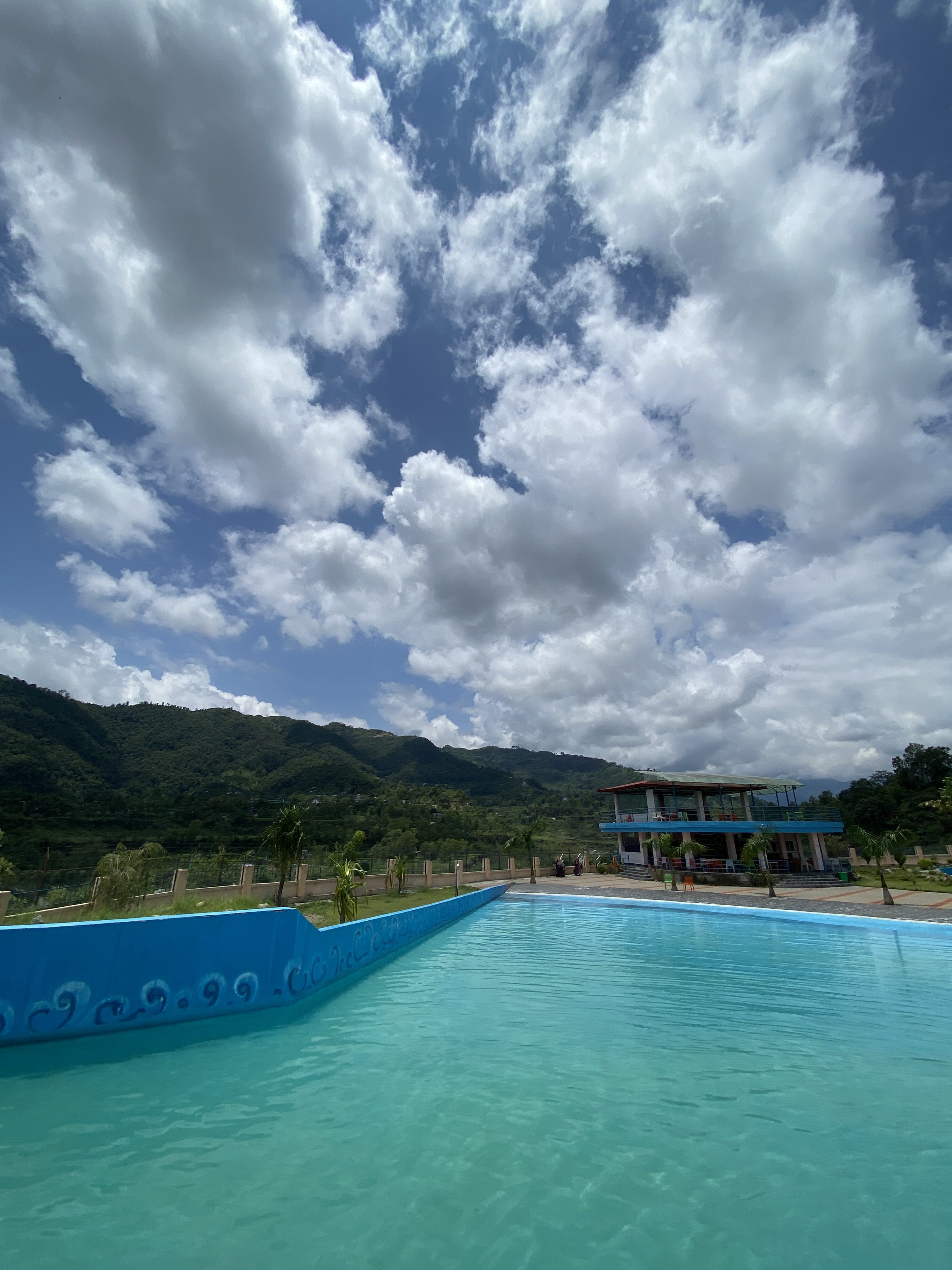 A scenic outdoor view featuring a large, clear blue swimming pool surrounded by lush green mountains under a bright sky filled with fluffy clouds.