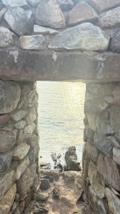 A weathered stone archway frames a view of the calm sea, with sunlight reflecting off the water's surface.