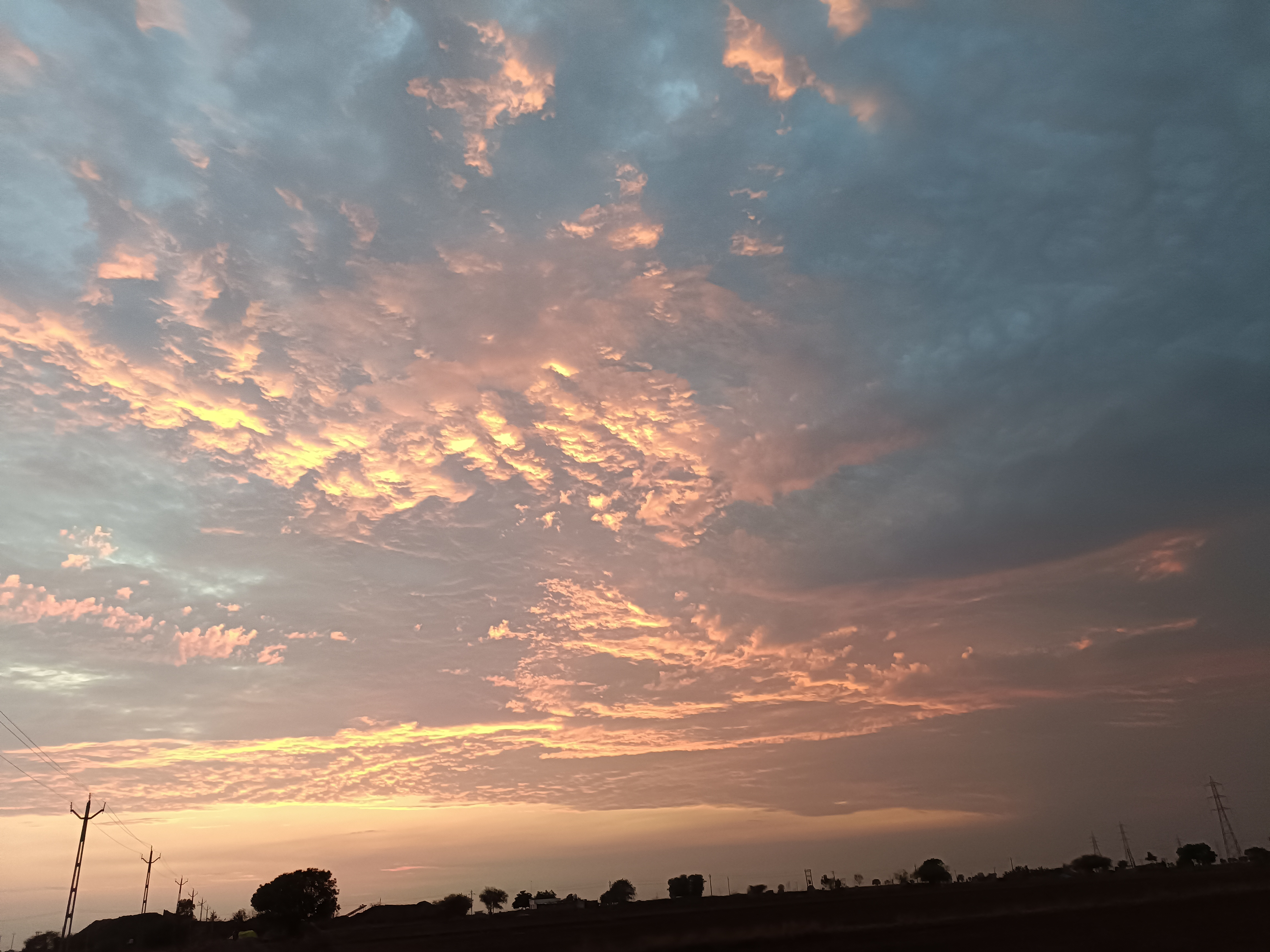 A dramatic sky at dusk, showcasing a blend of grey and pink clouds. The horizon glows with warm hues, while silhouettes of trees and power lines can be seen below, creating a serene landscape scene..