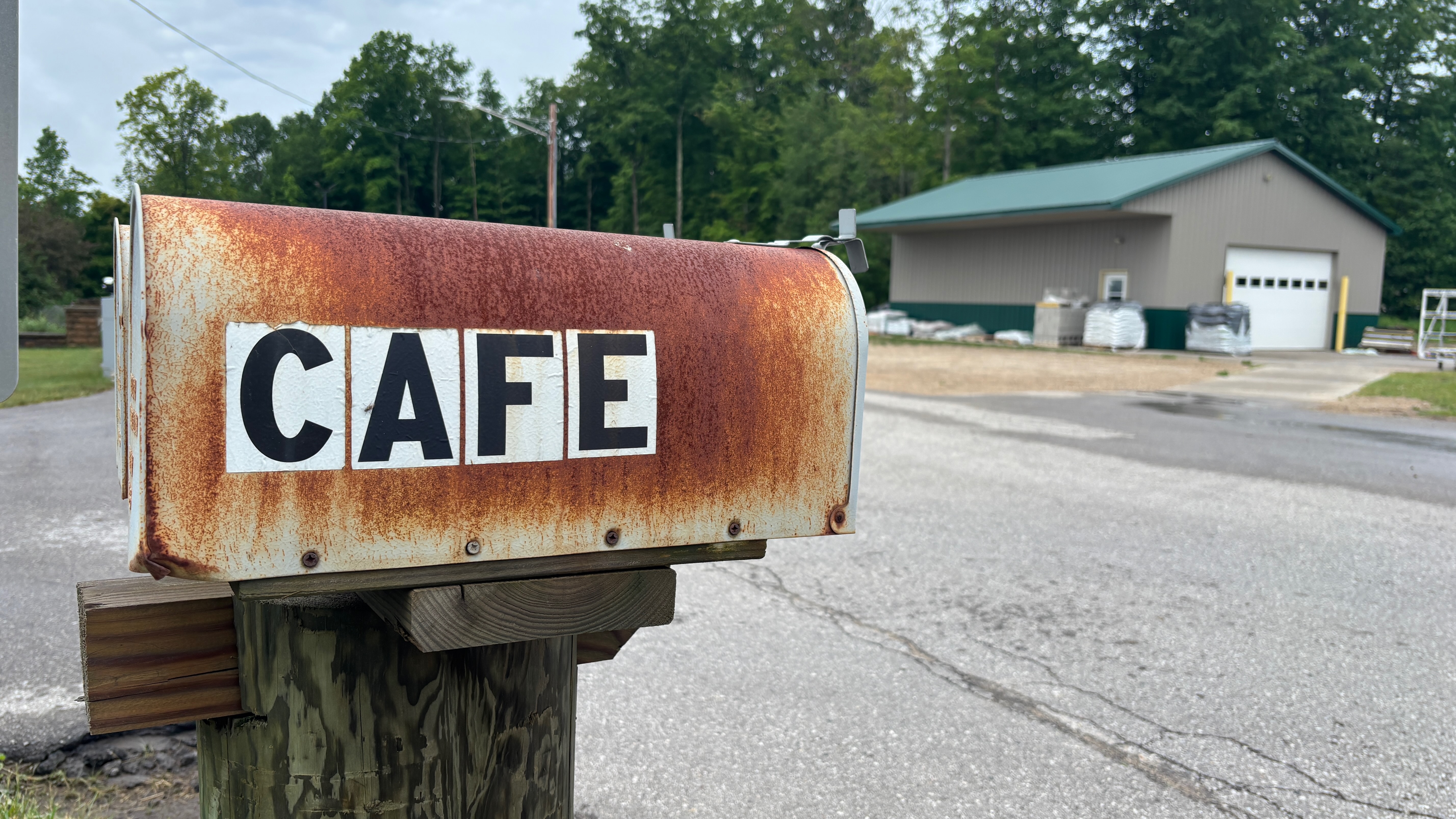 A rusty old mailbox with the word CAFE on it. A barn in the background.