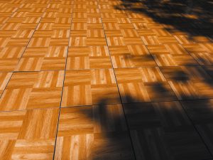 Polished wooden parquet flooring arranged in a basketweave pattern with alternating grain directions creates geometric squares. Dramatic shadows from trees streak diagonally across the warm honey-colored wood surface, creating stark contrast between illuminated and darkened sections of the floor.