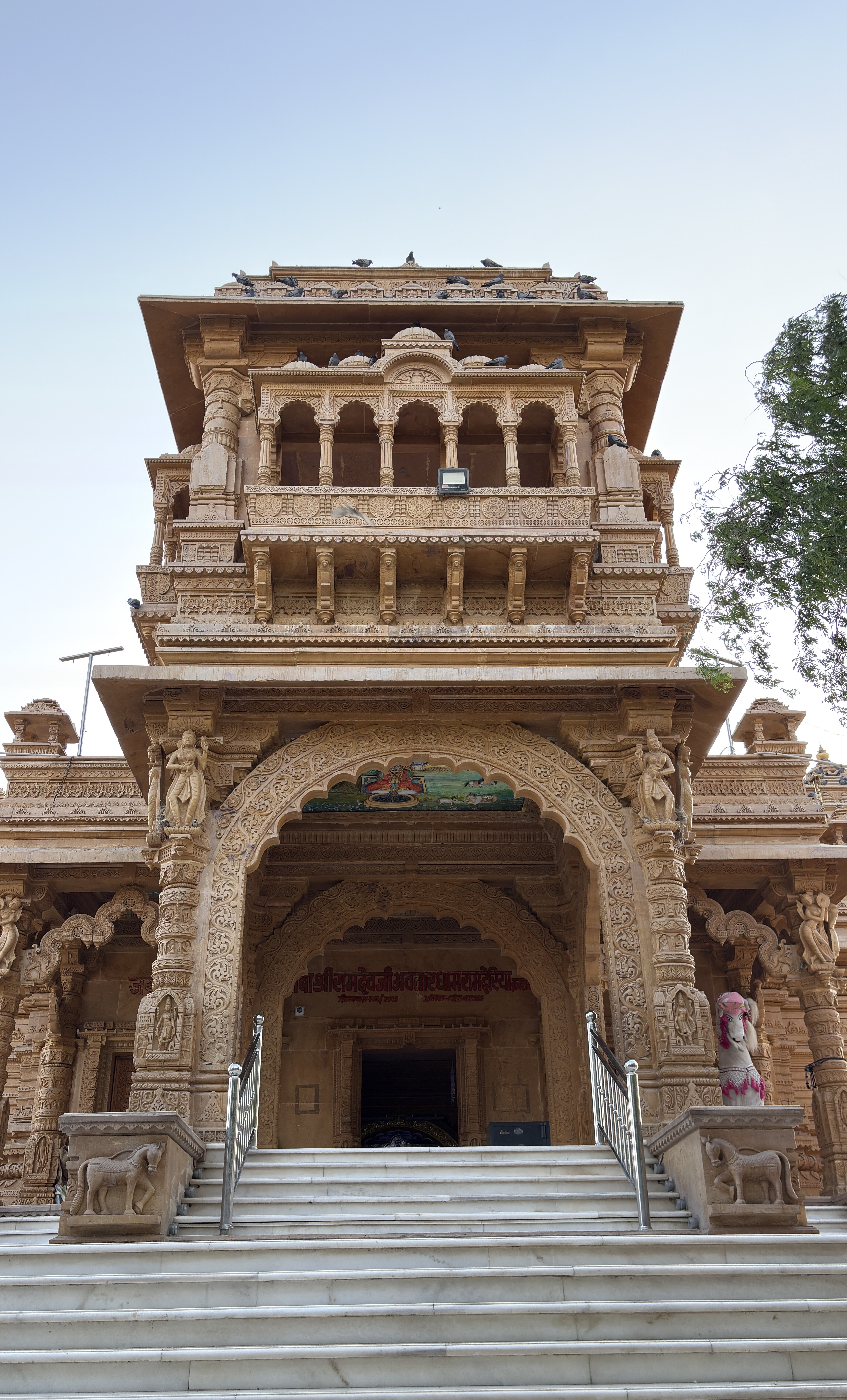Front view of an intricately carved sandstone temple entrance with marble steps, ornate pillars, and detailed sculptures, including divine figures and decorative motifs, under a clear sky.