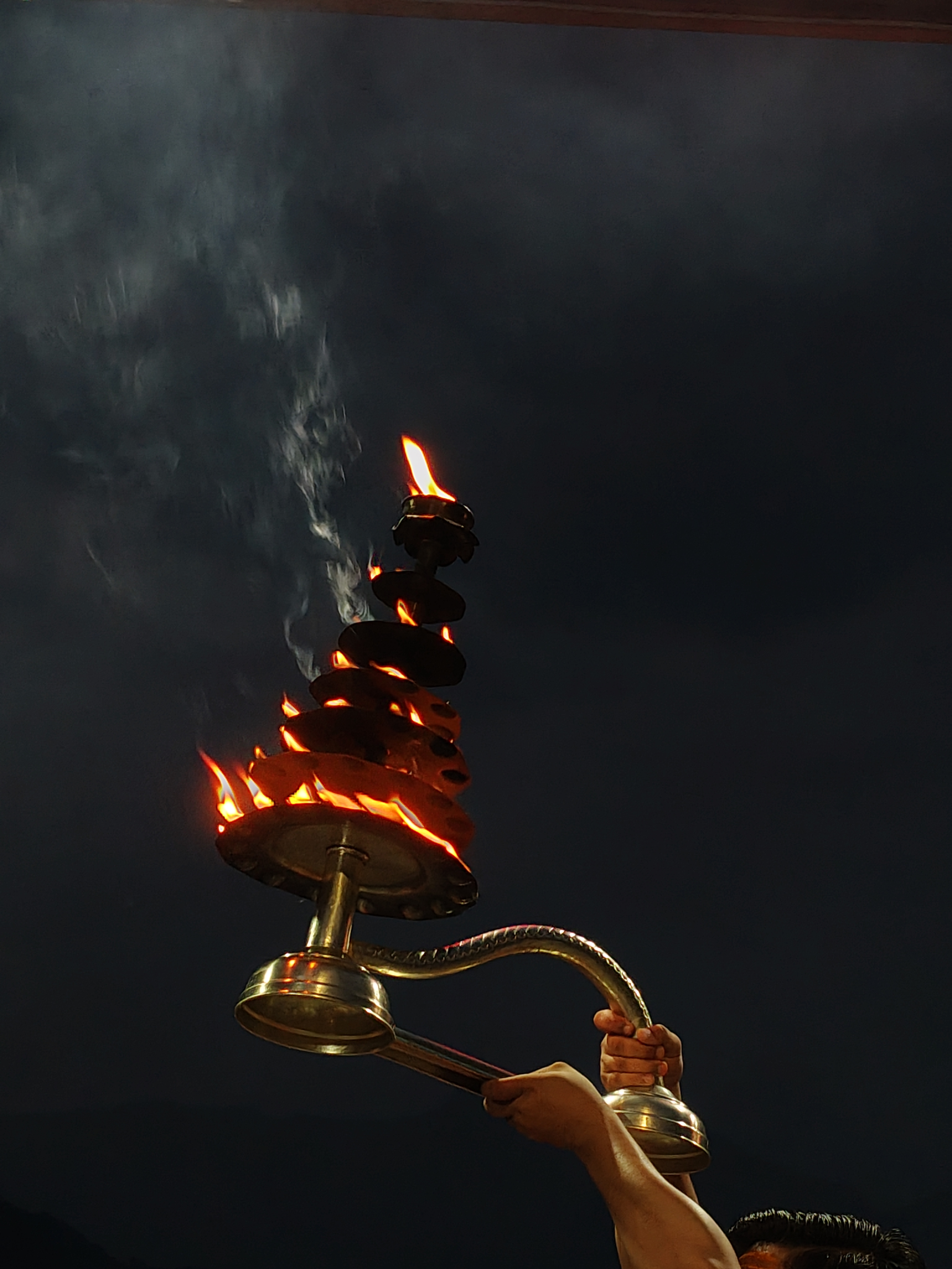 A person holding a decorative oil lamp with multiple flames burning brightly at the top, surrounded by dark, cloudy skies.