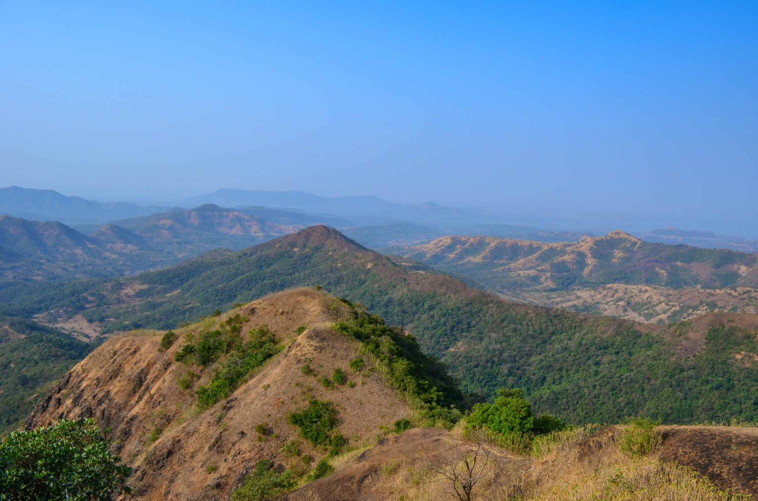 The landscape is a mix of dry, grassy patches and lush green vegetation, showcasing a mountainous terrain with varying elevations and contours. Distant hills are visible in the background, adding depth to the scene.