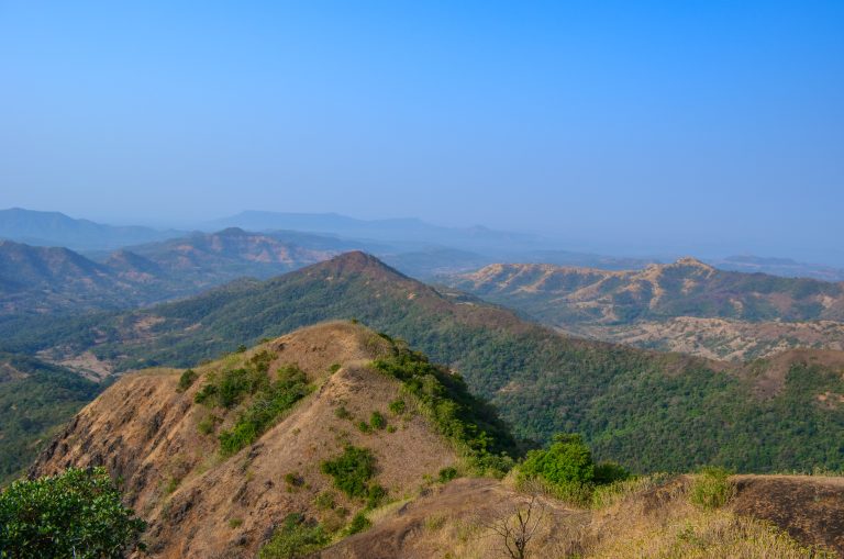 The landscape is a mix of dry, grassy patches and lush green vegetation, showcasing a mountainous terrain with varying elevations and contours. Distant hills are visible in the background, adding depth to the scene.