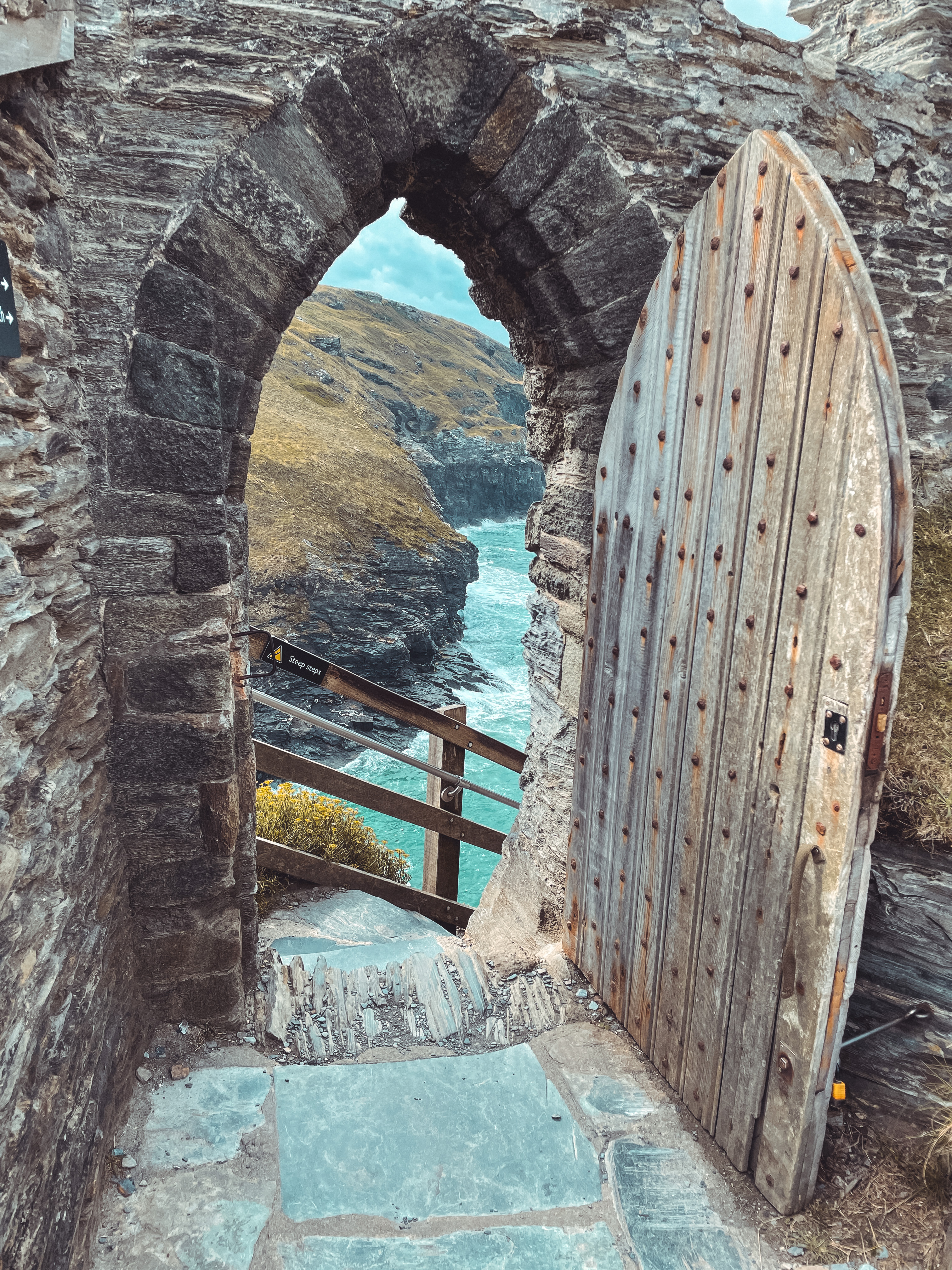 An old, weathered stone castle door stands open within crumbling ruins, revealing a dramatic view of rugged cliffs and the expansive sea beyond.
