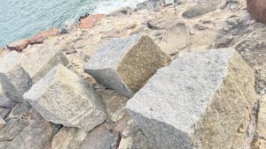Three large, weathered stone blocks are positioned on a rocky shoreline with the ocean visible in the background.